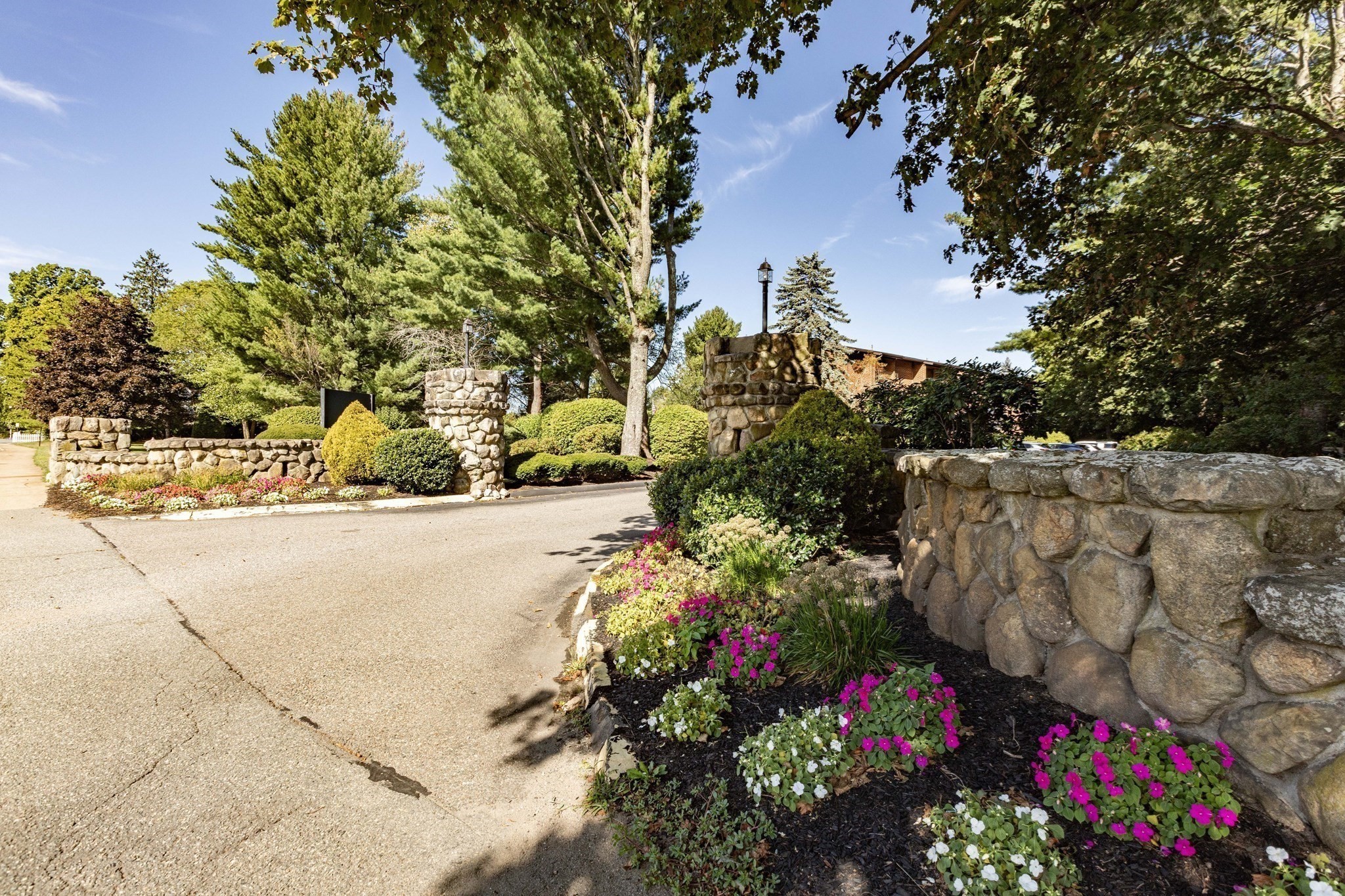 Residential entrance with stone walls, mature trees, and colorful flower beds along the driveway.