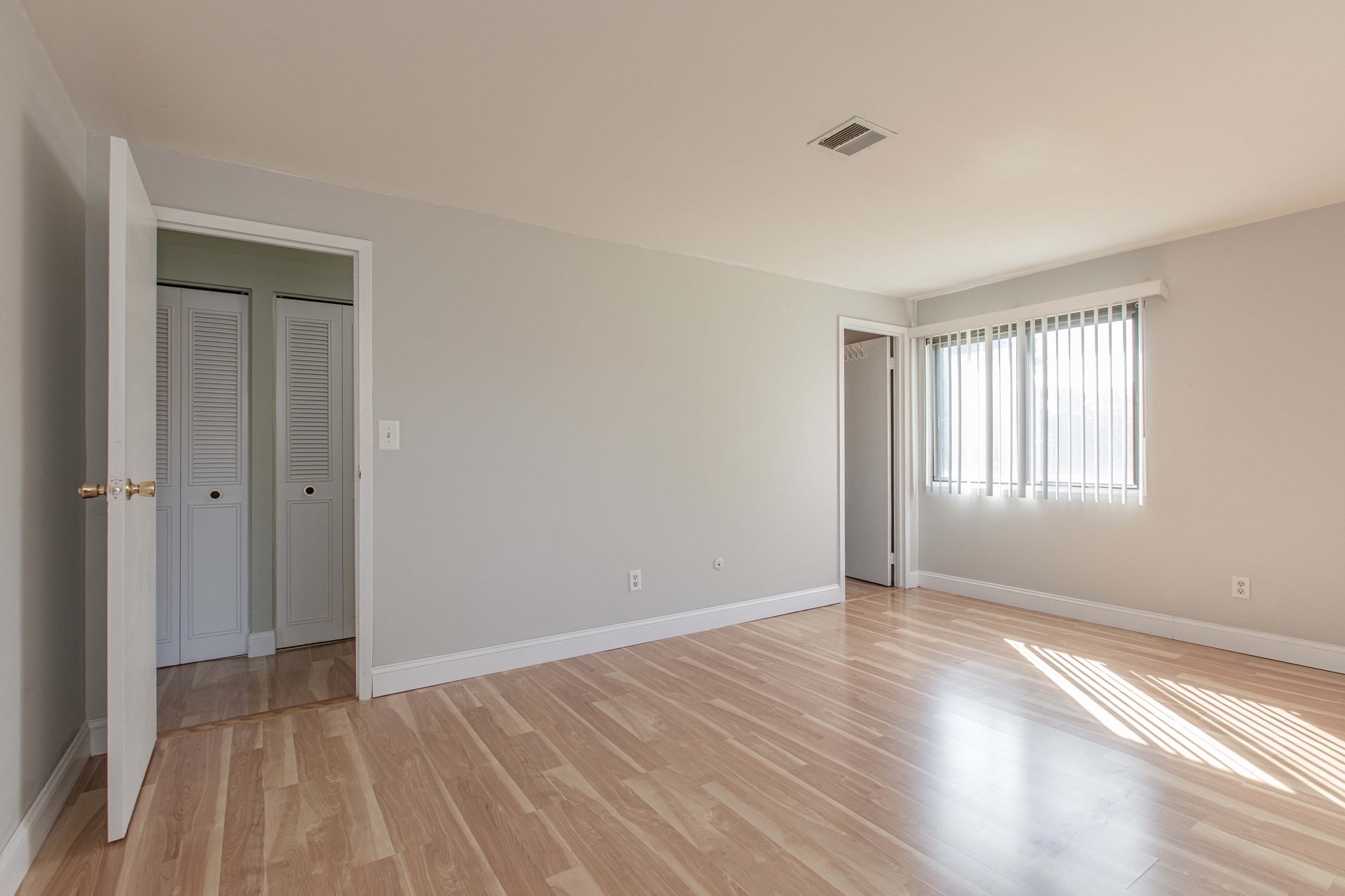 Empty bedroom with light wood floors and sunlight through vertical blinds.