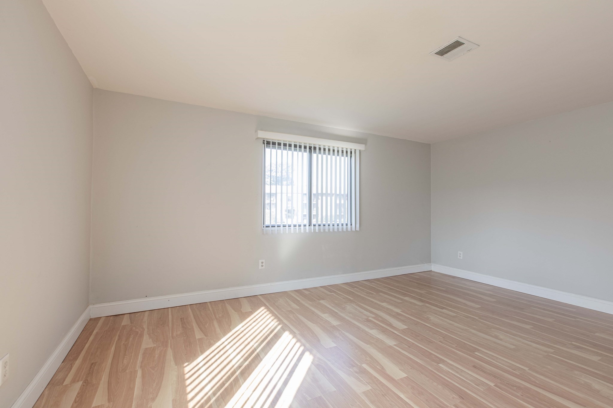 Empty room with wood-look flooring and sunlight streaming through vertical blinds.