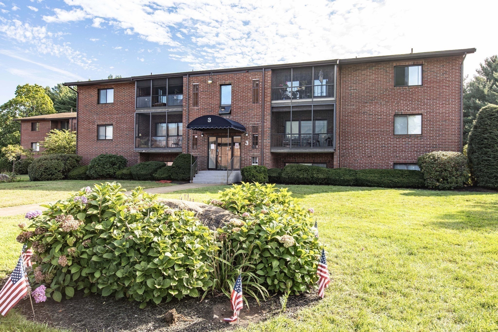 Red brick apartment building with screened balconies, a central entrance, and landscaped flower beds with American flags.