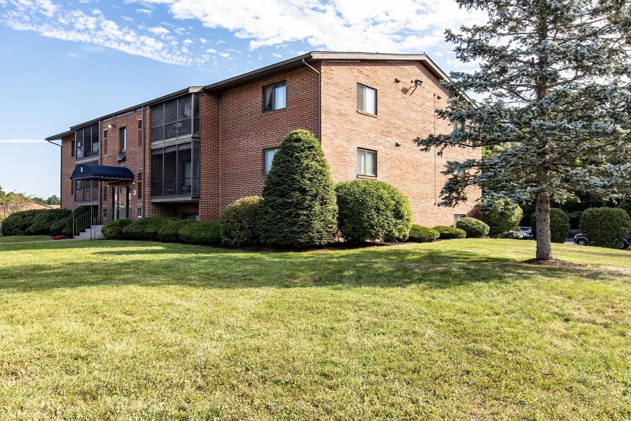 Red-brick apartment building with screened balconies, landscaped lawn, and a blue entrance awning marked 3.