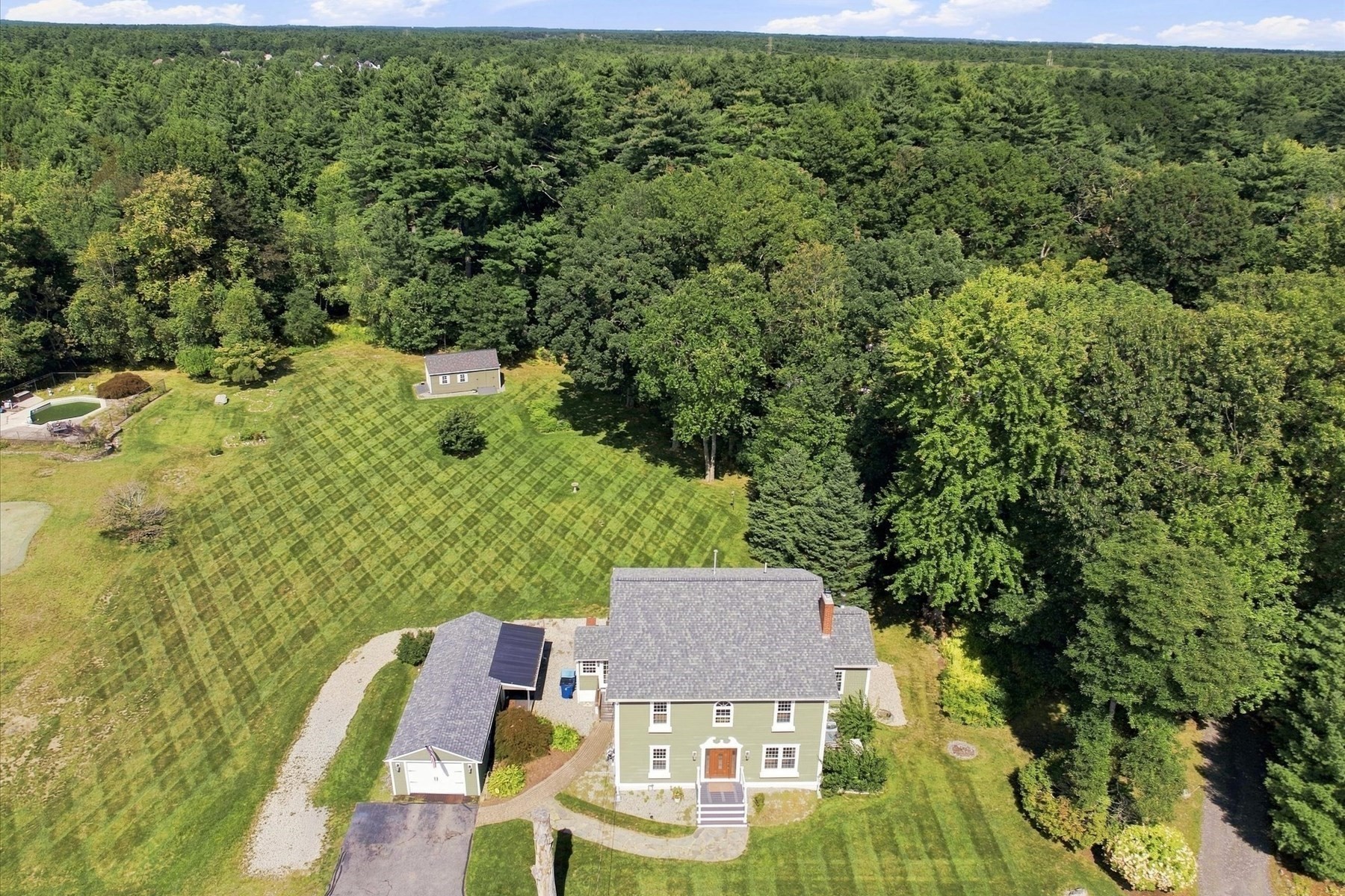 Aerial view of a two-story home, detached outbuilding, expansive lawn, pool, and surrounding woodland.