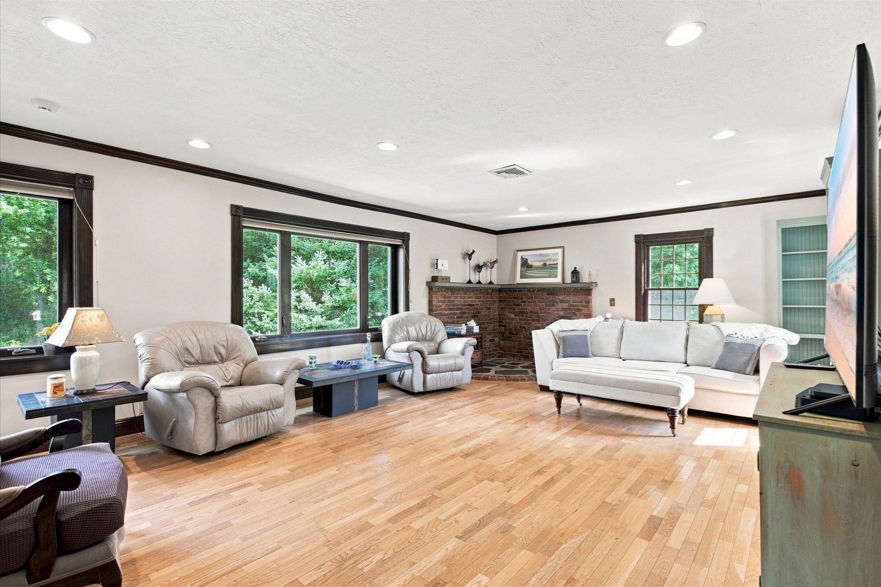 Living room with hardwood floors, brick fireplace, large windows, leather armchairs, and a white sectional sofa.