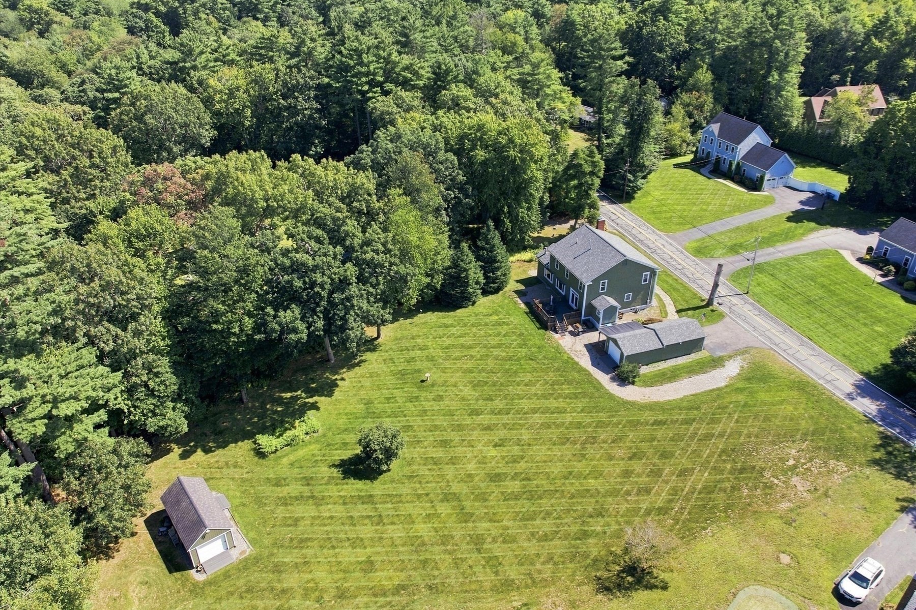 Aerial view of a green house and detached garage on a broad lawn beside dense woods.