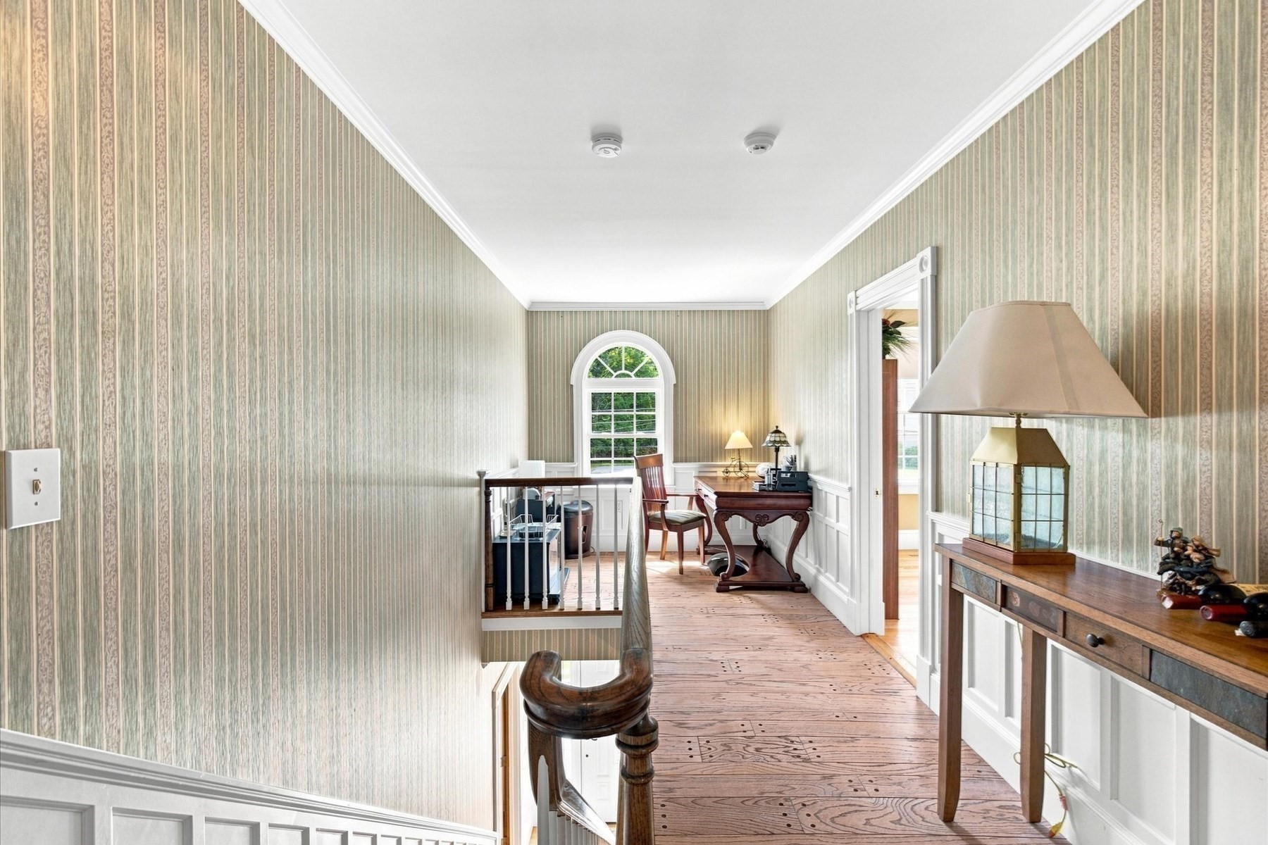 Upstairs hallway with striped wallpaper, wide-plank wood flooring, and an arched window.
