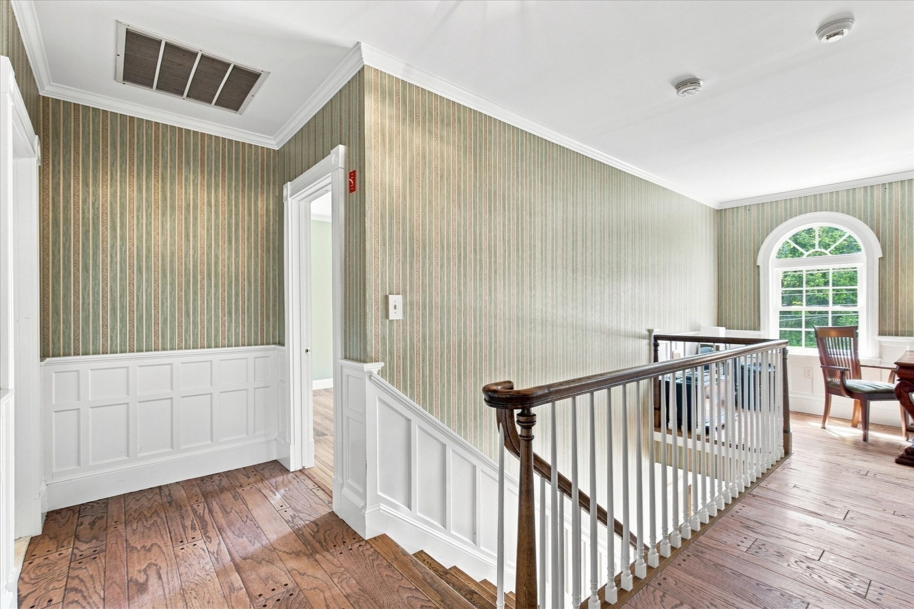 Landing with striped wallpaper, paneled wainscoting, wood floors, and an arched window.