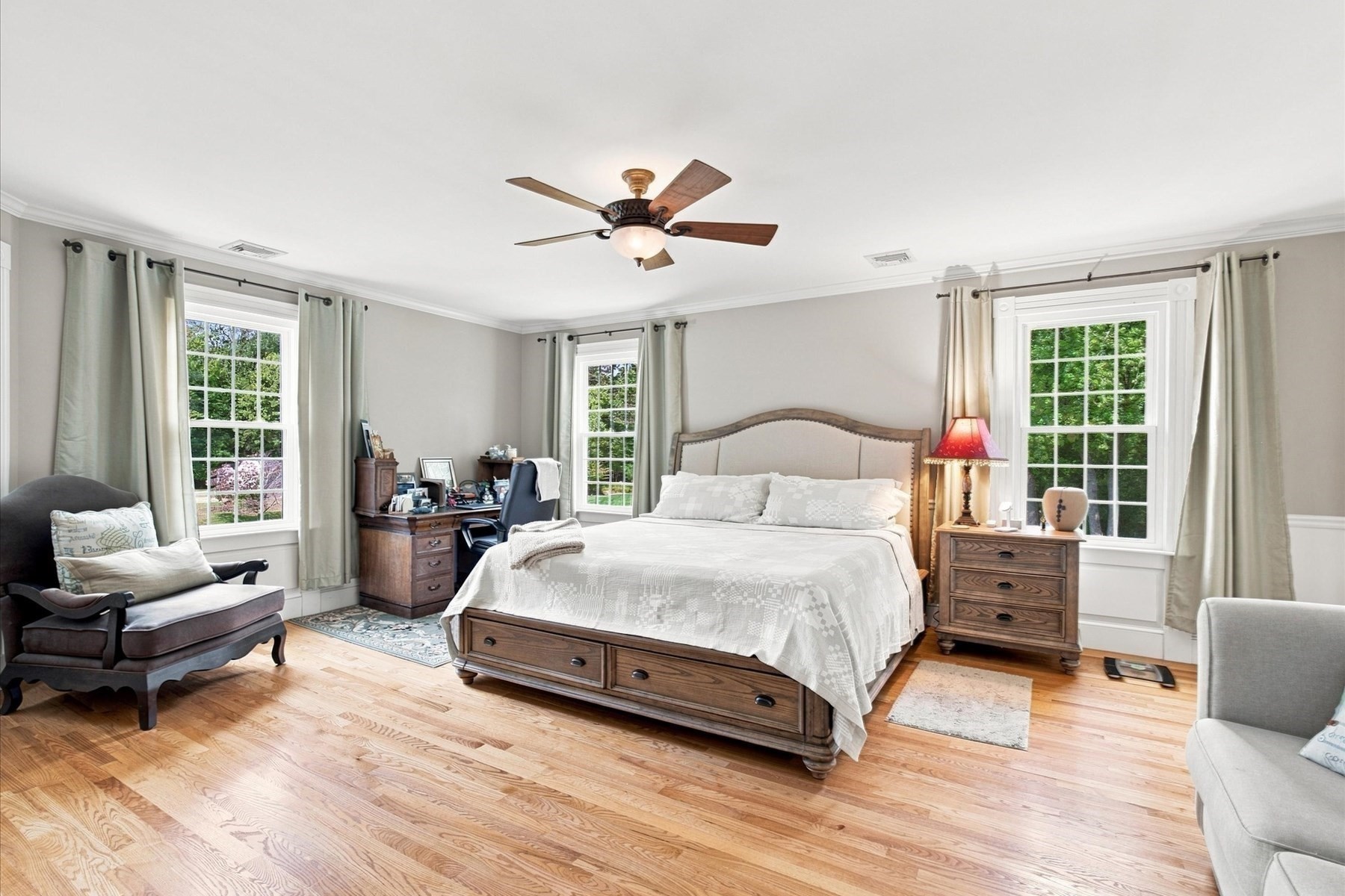 Bedroom with a large wooden bed, white bedding, hardwood floors, and three curtained windows.