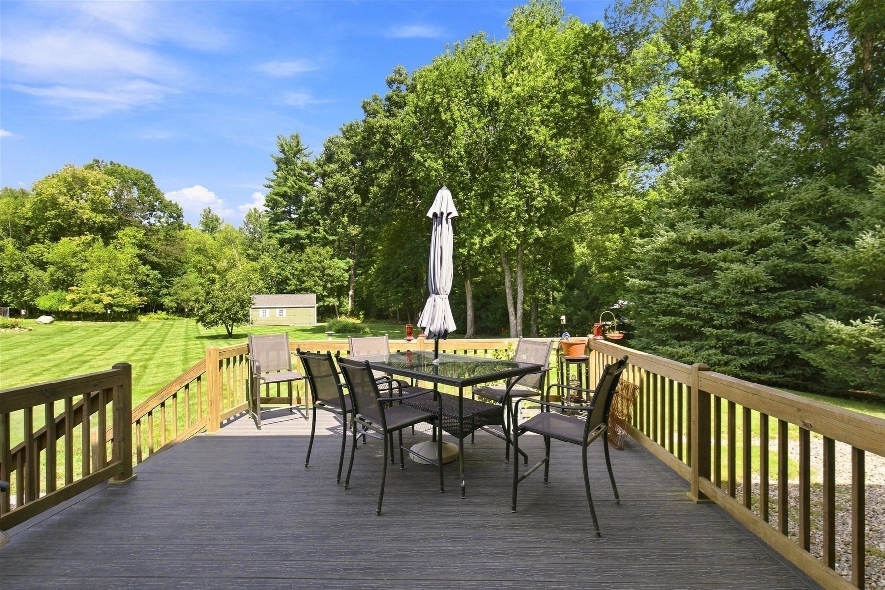 Wooden deck with outdoor dining set overlooking a spacious lawn bordered by mature trees.