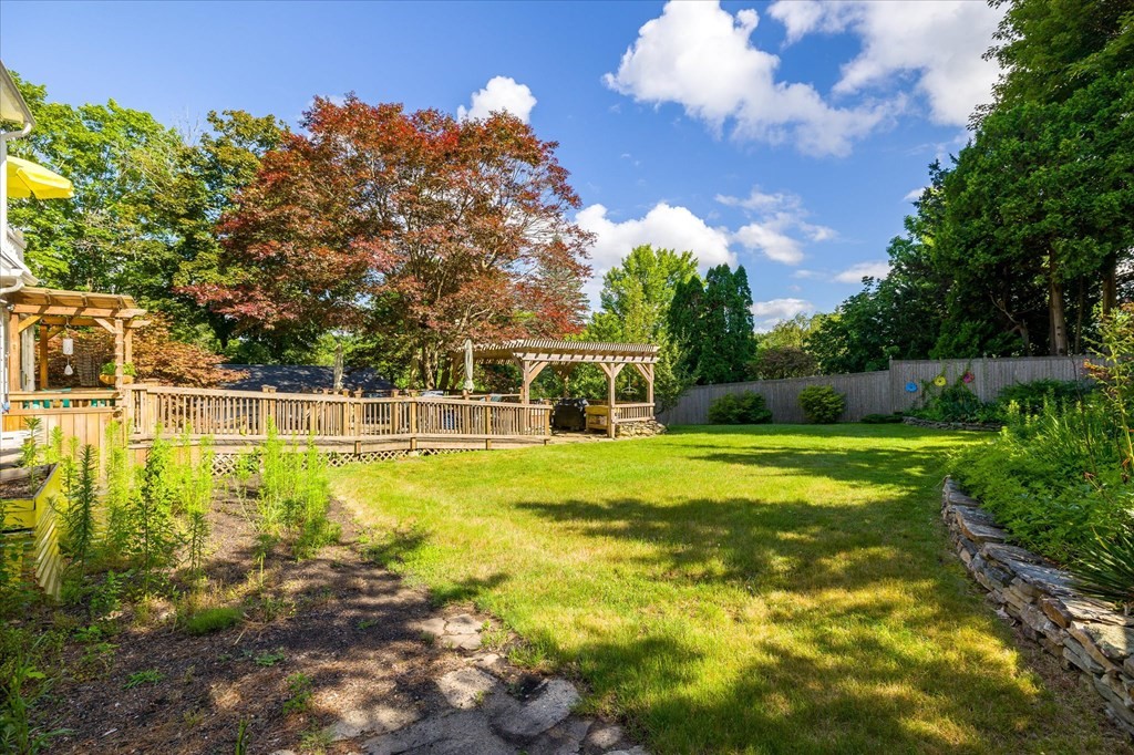 Backyard lawn bordered by mature trees, with a wooden deck, pergola, and privacy fence.