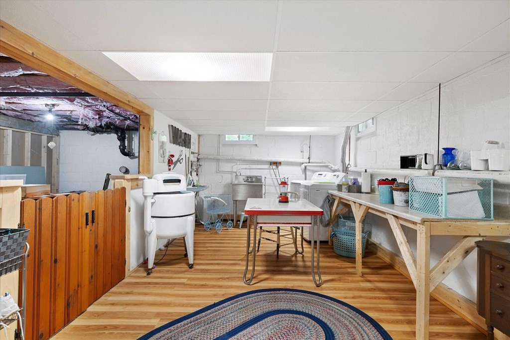 Basement laundry room with washing machines, utility sink, and worktables on wood-look flooring.