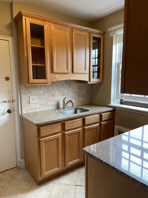 Kitchen with honey-colored cabinets, granite countertops, tiled backsplash, and a sink beneath a window.
