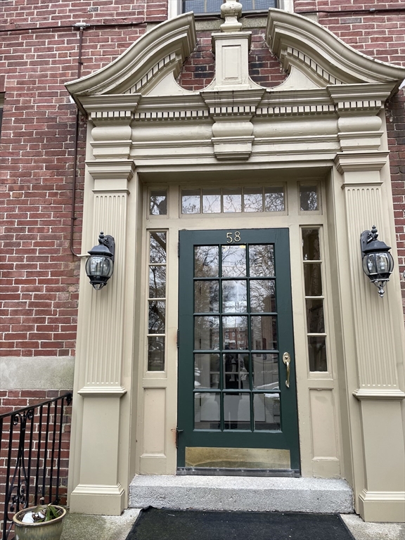 Ornate brick building entrance with cream trim, paired lanterns, and a dark green glass-paneled door.