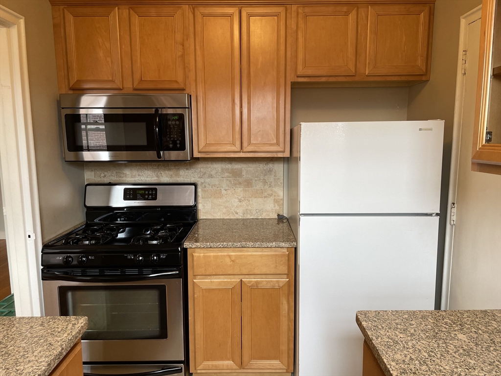 Kitchen with honey-colored cabinets, a gas range, over-the-range microwave, white refrigerator, and granite counters.
