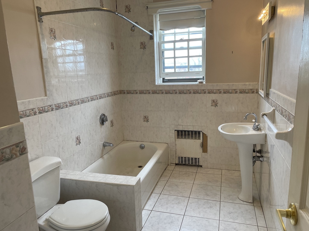 Tiled bathroom with a bathtub, pedestal sink, toilet, and window above a wall heater.