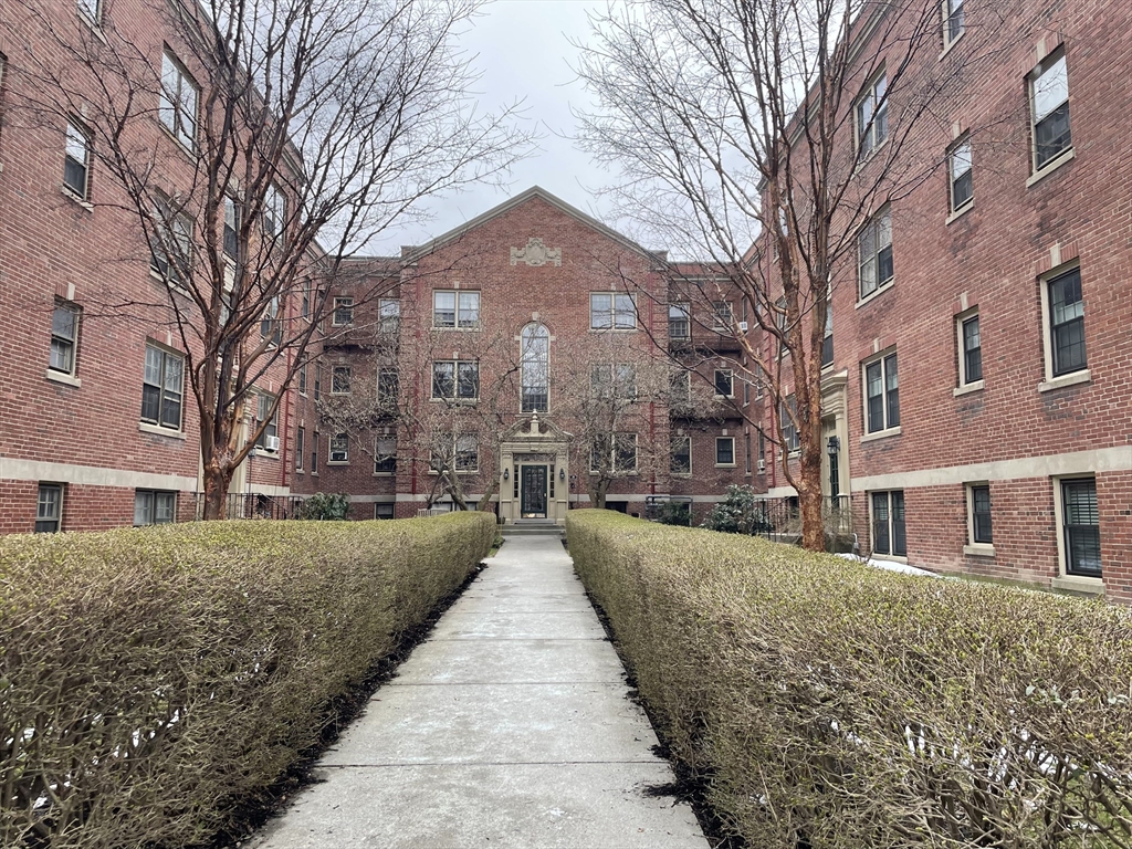 Red-brick apartment building with a central entrance, framed by hedges and leafless trees.