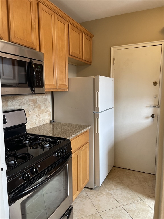 Narrow kitchen with wood cabinets, black gas range, white refrigerator, and entry door at the far end.