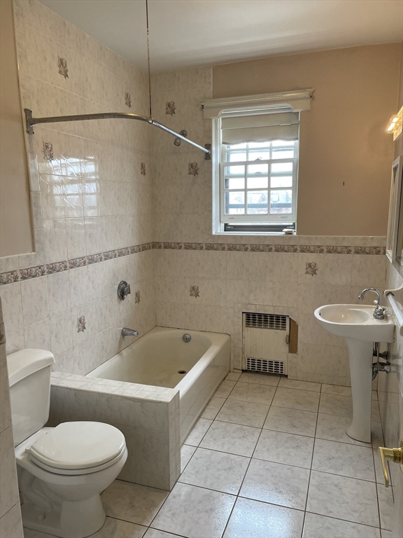 Bathroom with cream patterned tile, built-in tub, pedestal sink, and window above a wall heater.