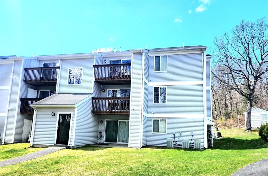 Gray condominium building with dark balconies, a covered entrance, and a lawn.
