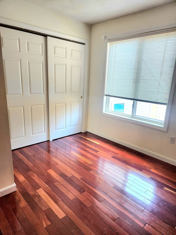 Empty bedroom with reddish-brown hardwood floors, sliding closet doors, and a window with blinds.