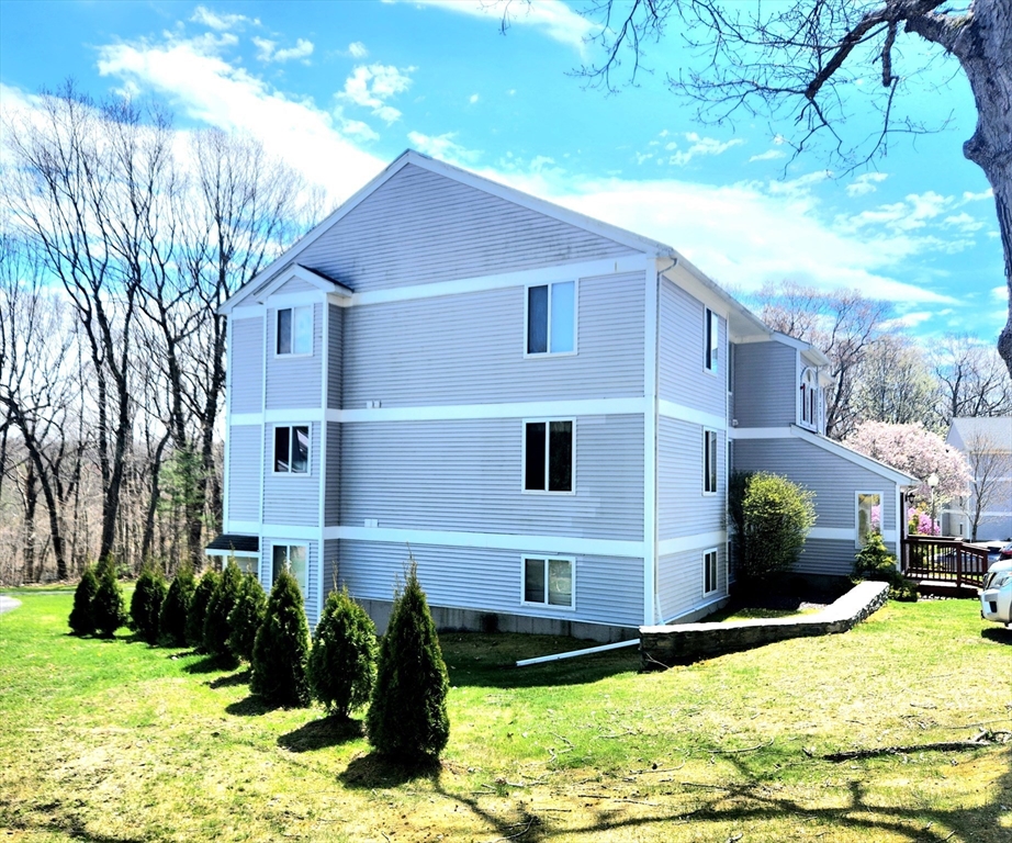 Three-story gray condominium building with white trim, evergreen shrubs, and a sunny lawn.