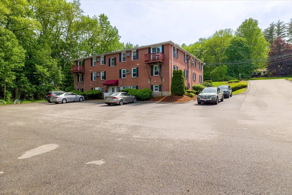 Three-story red brick apartment building with balconies, a red entrance awning, and cars in the parking lot.