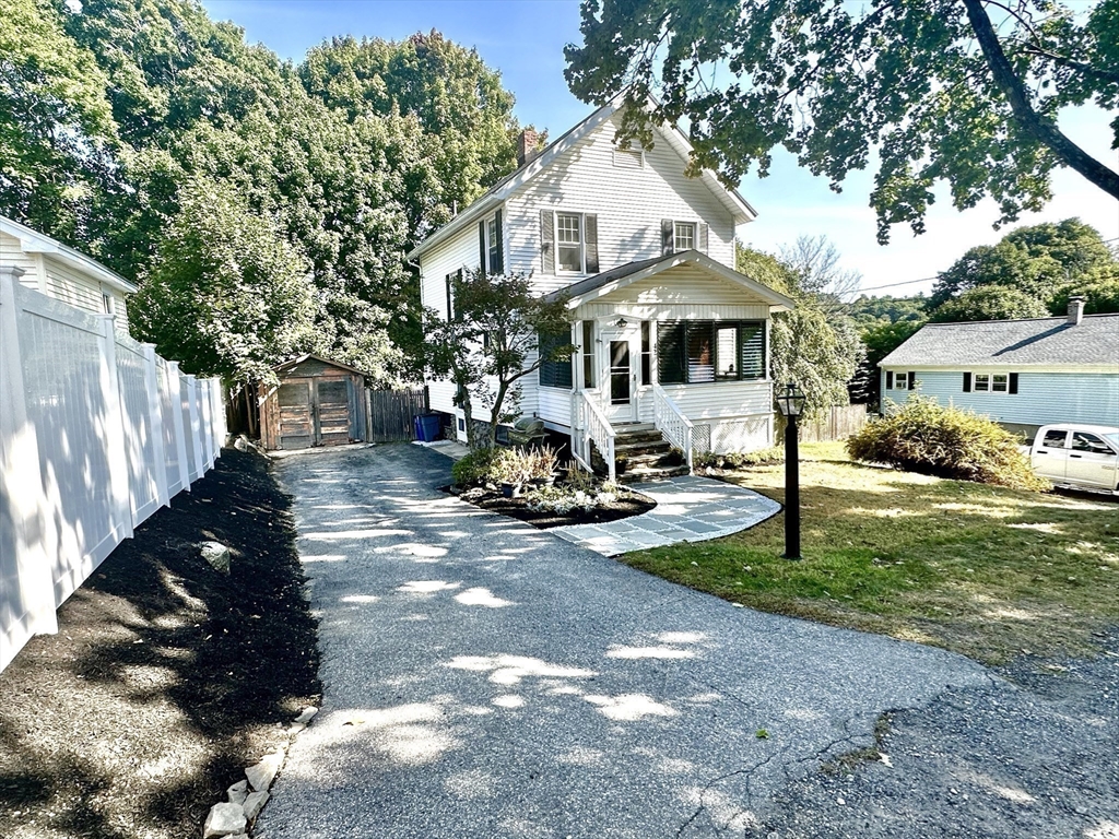 White two-story house with a covered porch, curved walkway, and driveway beside a wooden shed.