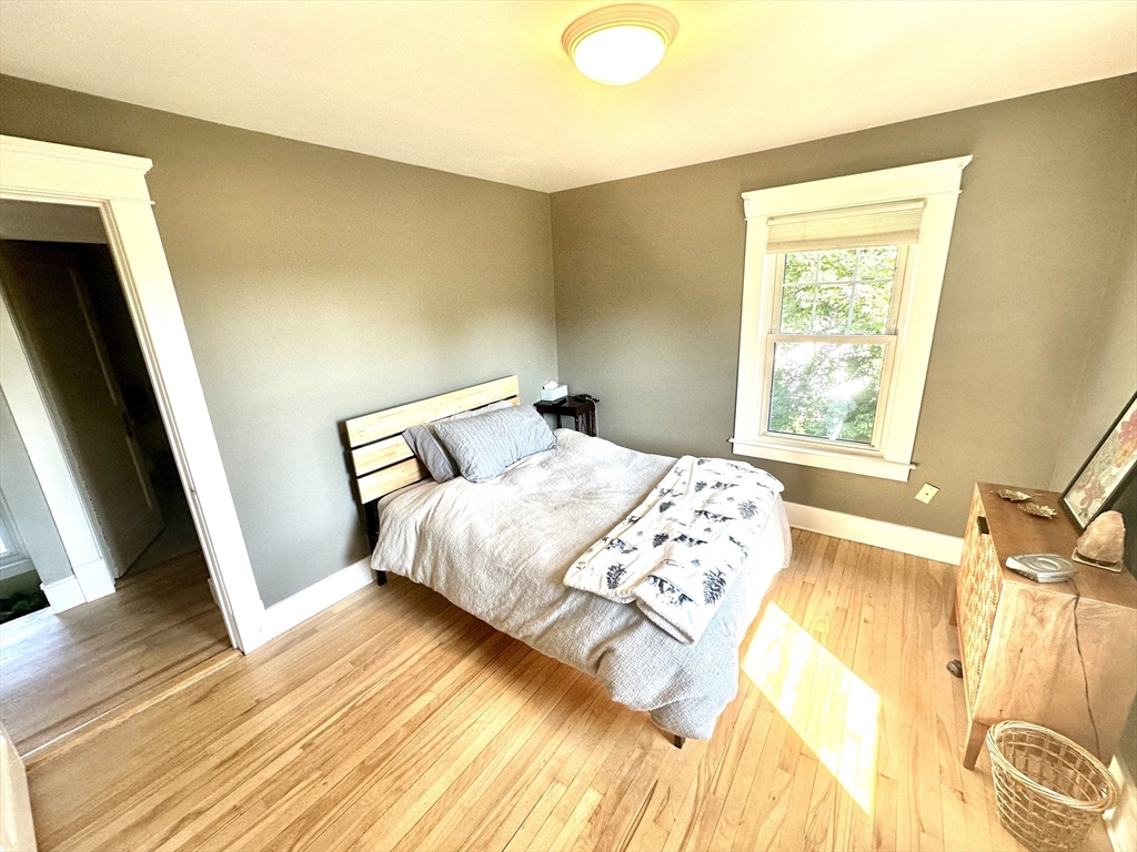 Bedroom with a bed, sunlit hardwood floors, and a white-trimmed window.