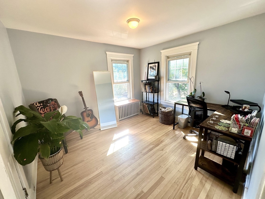 Home office with two tall windows, light wood flooring, a desk, guitar, and large potted plant.