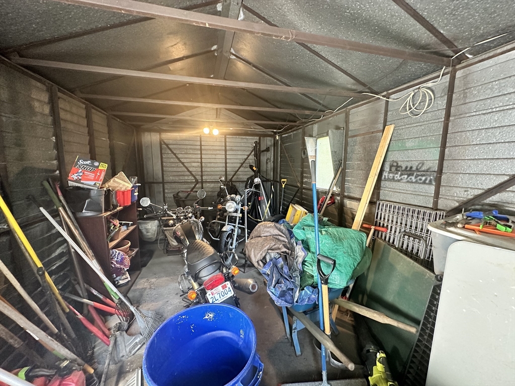 Cluttered storage shed with motorcycles, gardening tools, and a blue barrel.