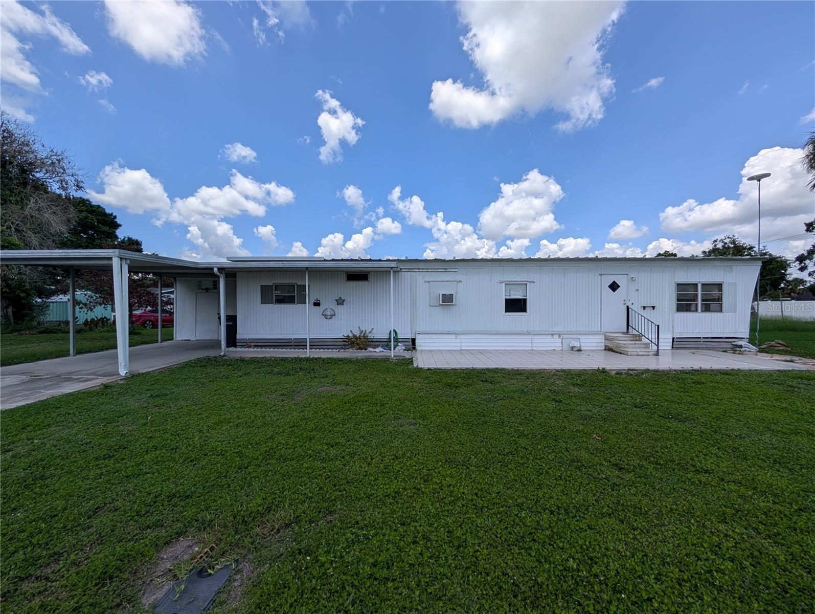 White manufactured home with a covered carport, tiled patio, and broad front lawn.