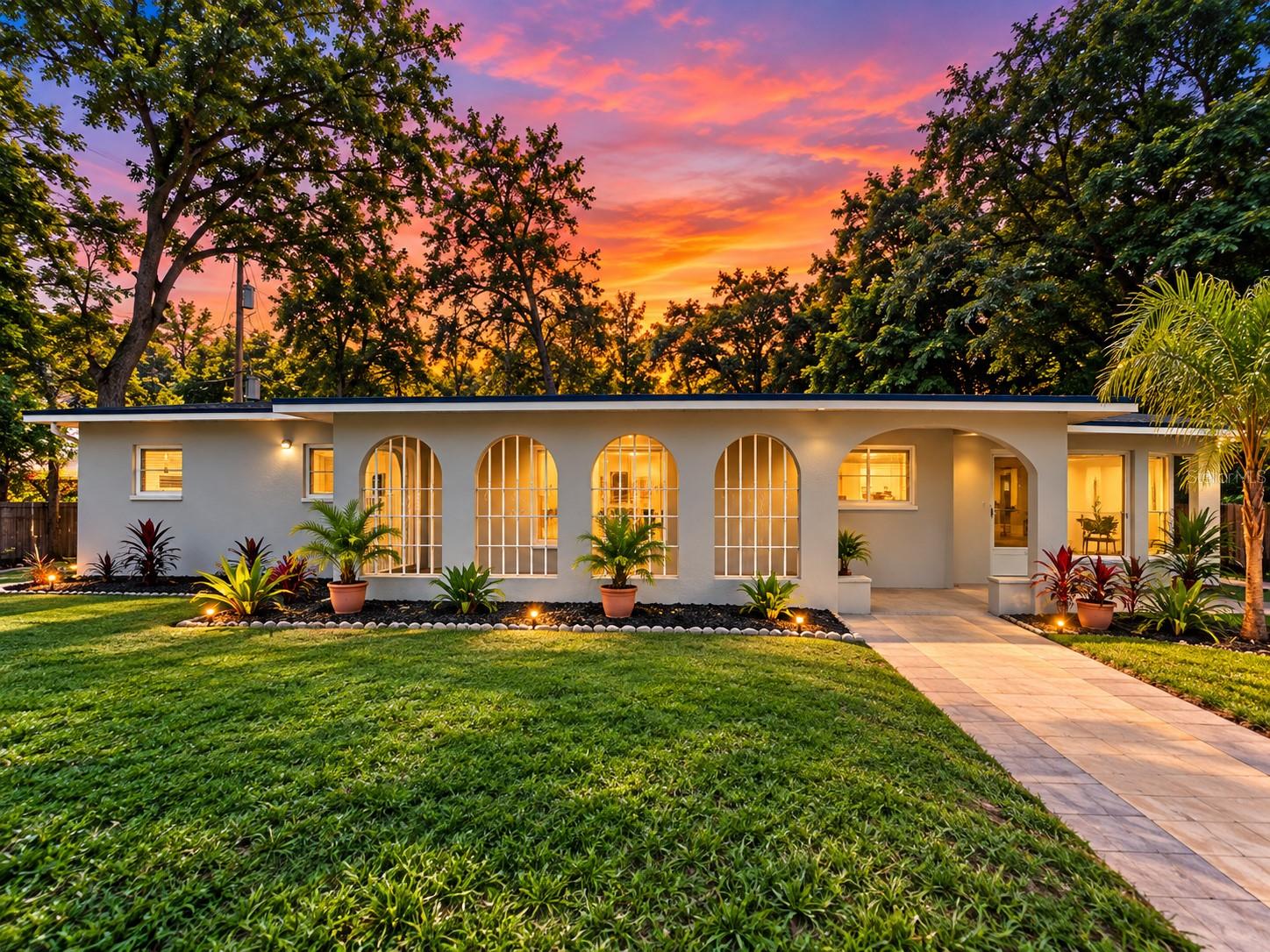 Single-story home with arched windows, tropical landscaping, and a paved front walkway at sunset.