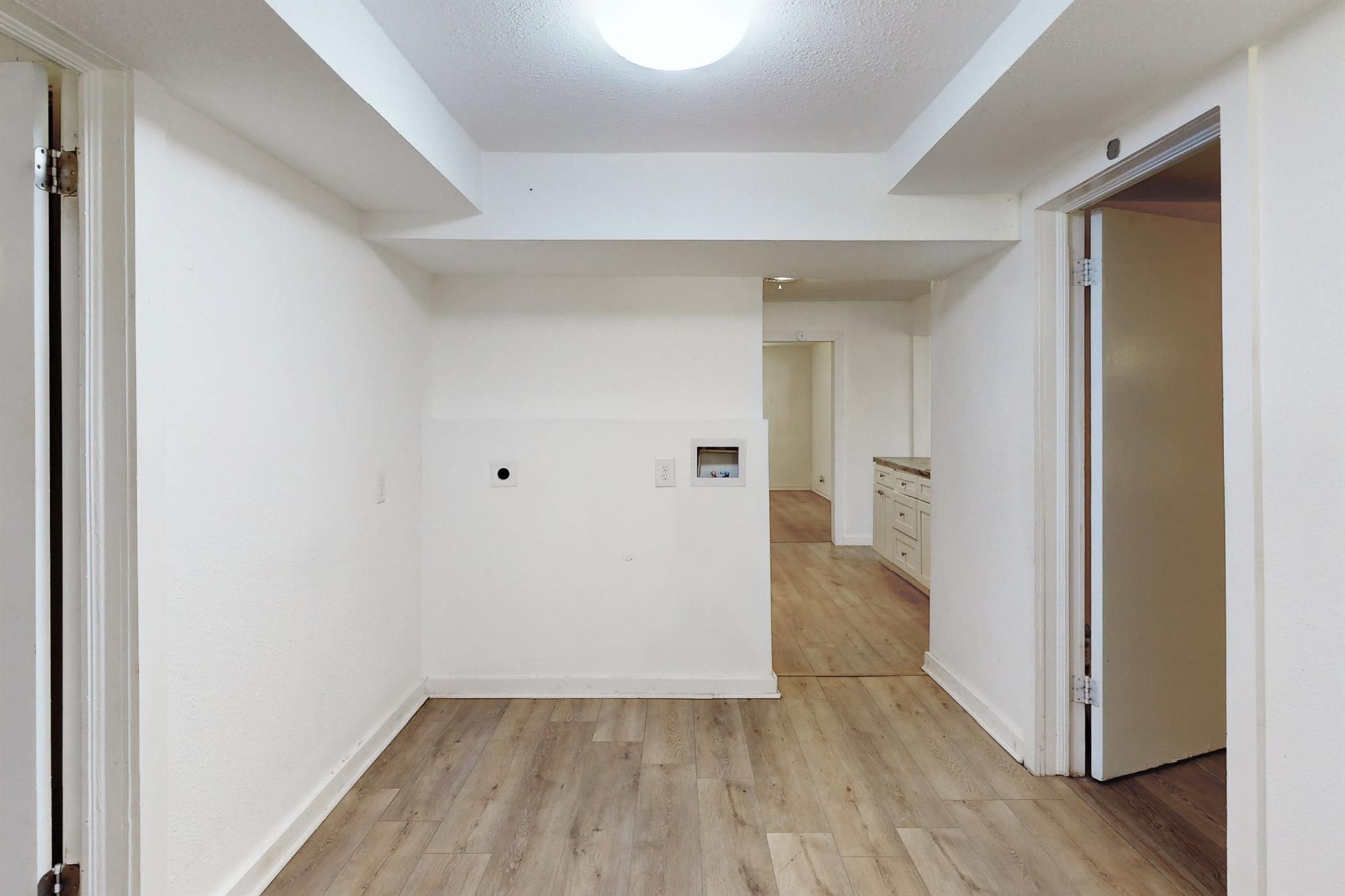 Empty room with wood-look floors, a utility connection, and kitchen cabinets visible beyond.