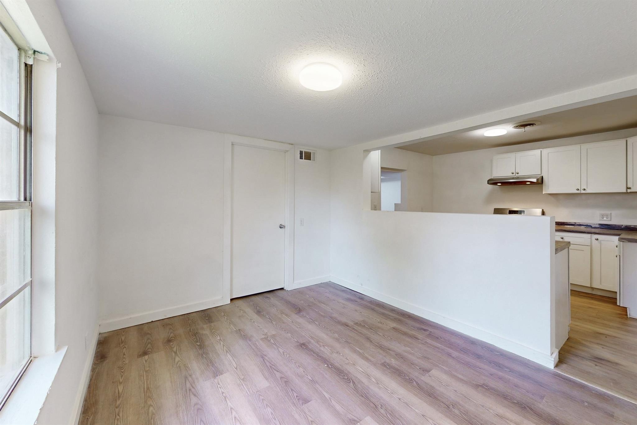 Unfurnished living area with wood-look flooring, a large window, and an open kitchen with white cabinets.