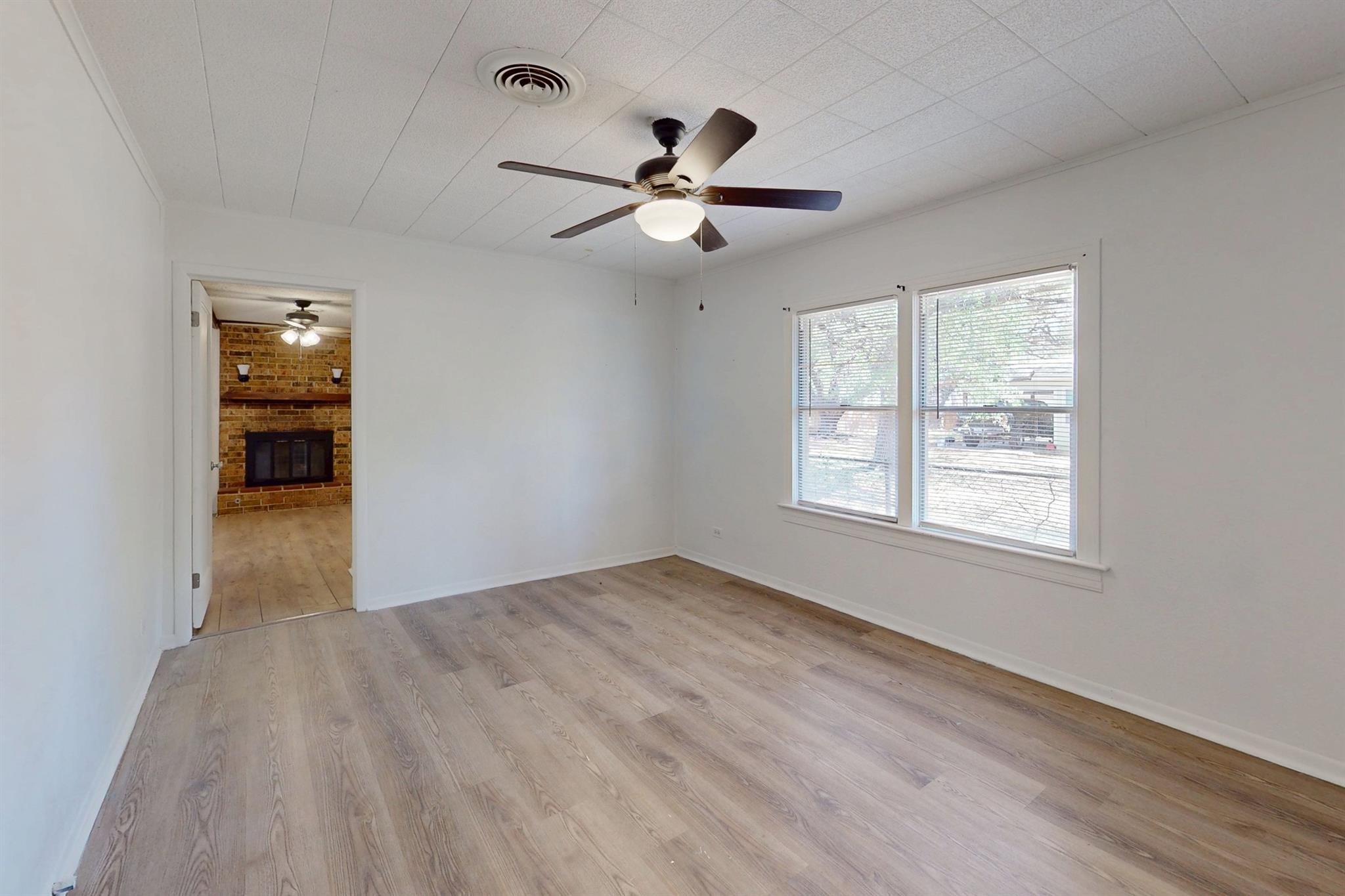 Empty room with wood-look flooring, ceiling fan, and windows; brick fireplace visible through the doorway.