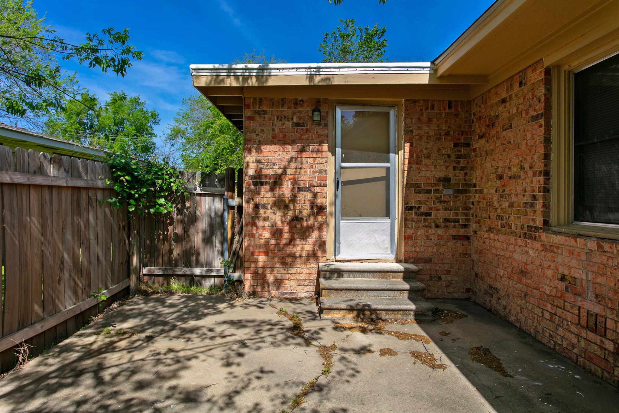 Concrete patio beside a brick house, with steps to a screened door and a wooden privacy fence.