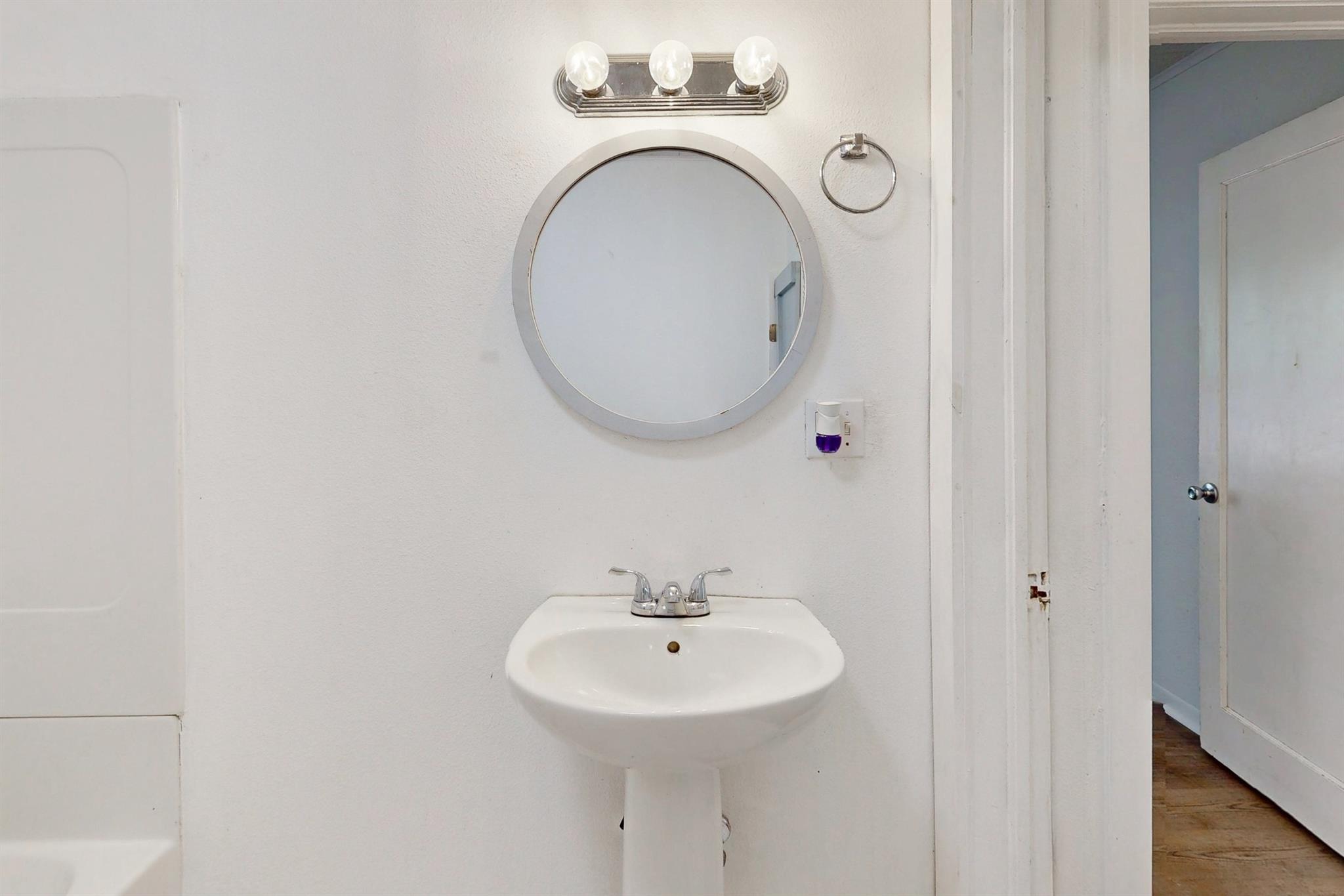 Bathroom with a white pedestal sink, round mirror, and three-bulb light fixture.
