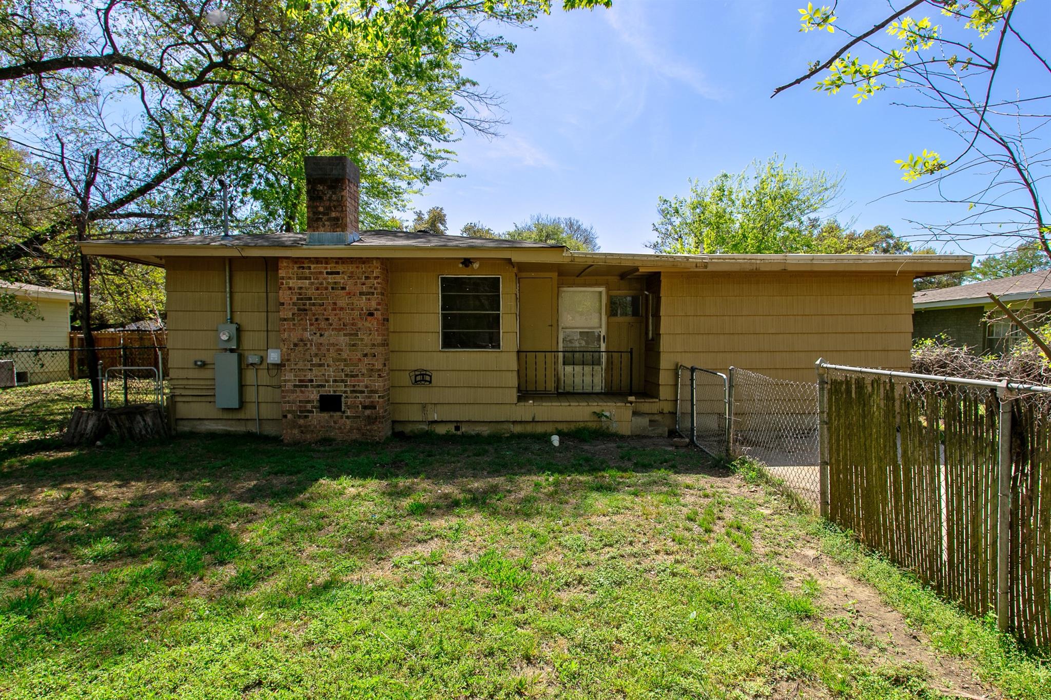 Rear of a single-story tan home with a brick chimney and fenced grassy backyard.
