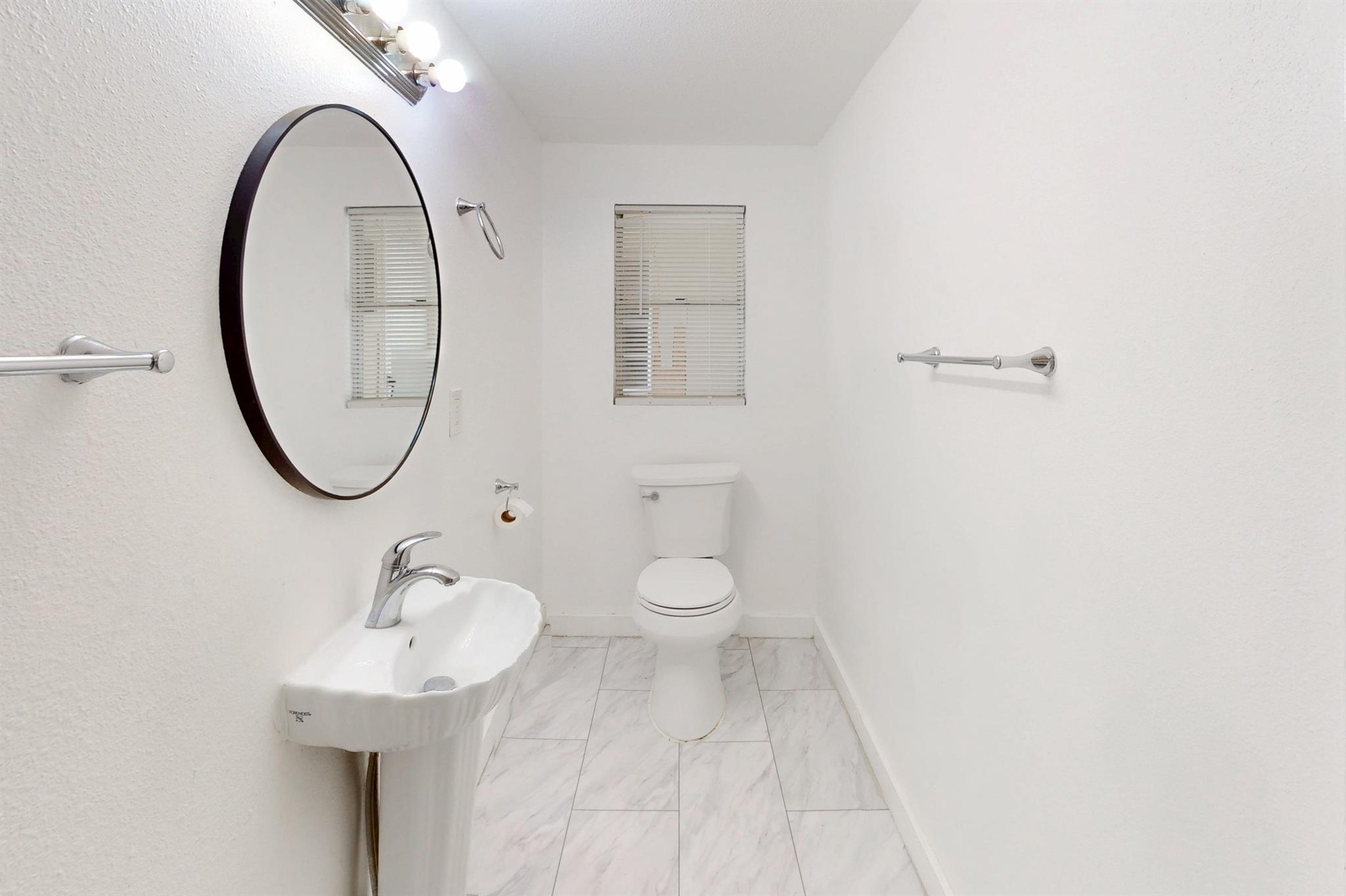 White bathroom with a pedestal sink, large round mirror, toilet, and marble-look tile flooring.