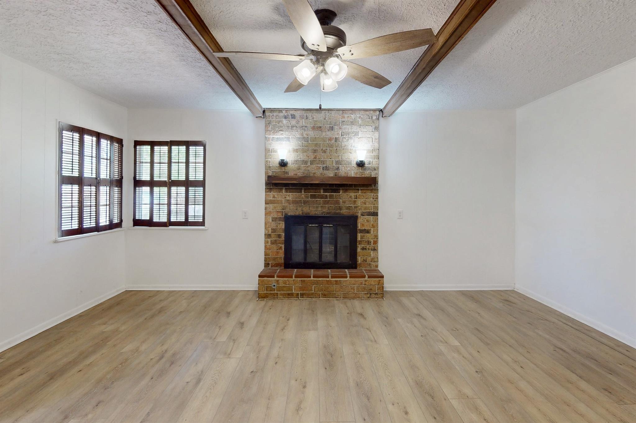 Empty living room with a brick fireplace, dark wood shutters, and exposed ceiling beams.