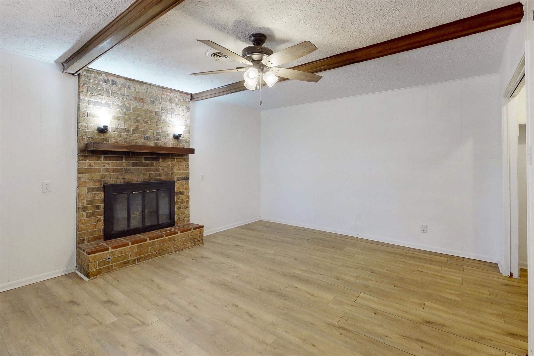 Empty living room with a brick fireplace, exposed ceiling beams, and light wood-look flooring.