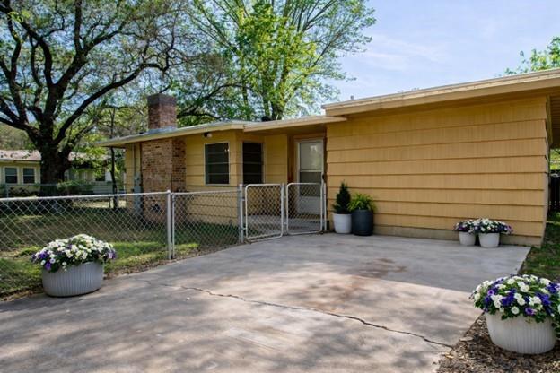 Single-story yellow house with a brick section, chain-link fence, and flower pots beside the concrete driveway.