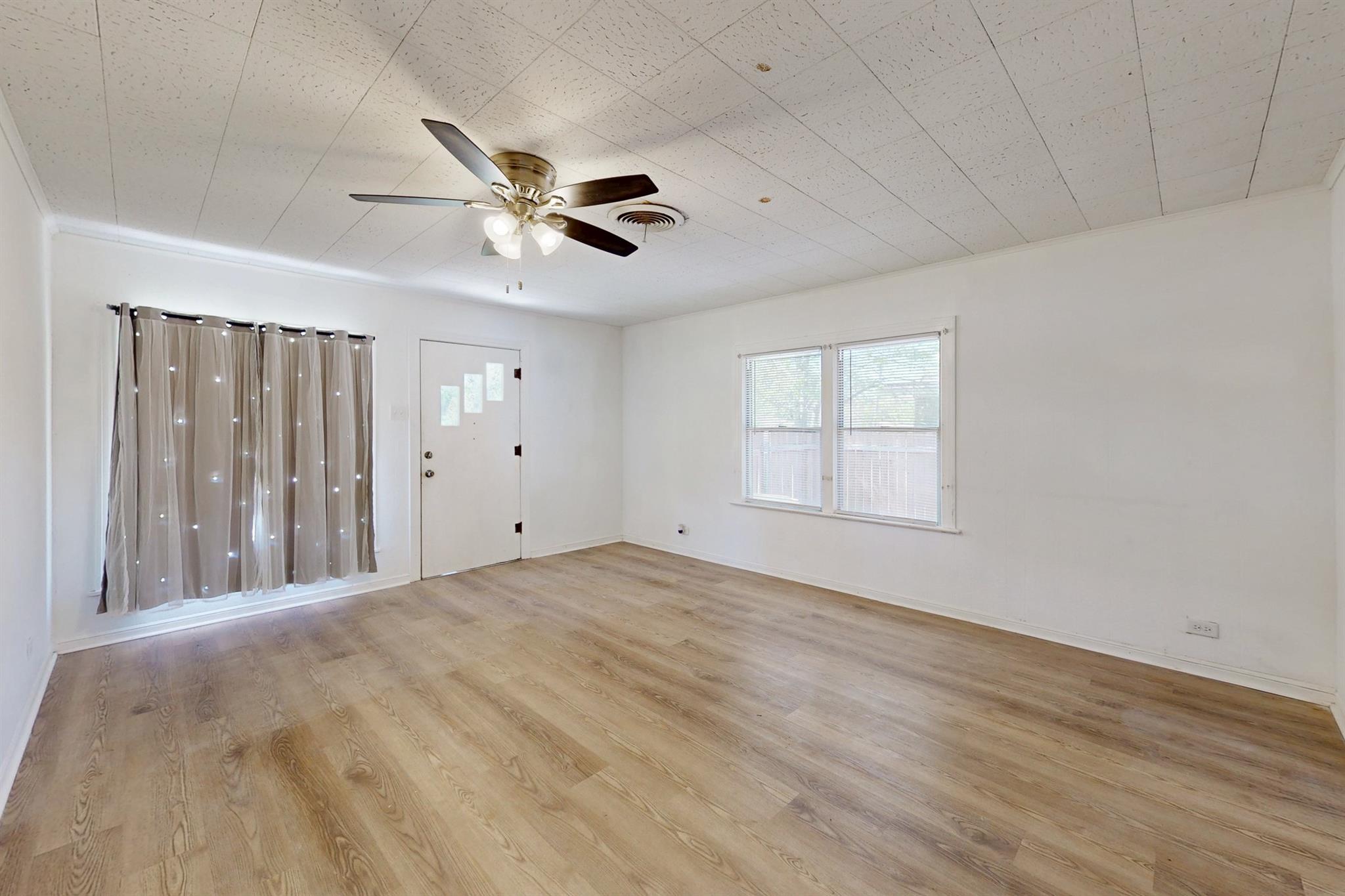 Empty room with light wood-look flooring, curtained glass sidelights, two windows, and a ceiling fan.