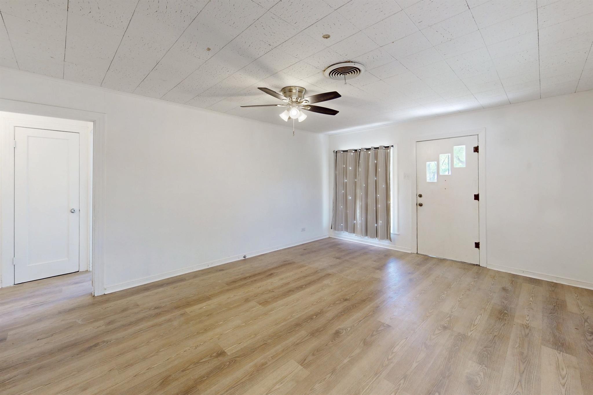 Empty room with wood-look floors, ceiling fan, and curtained window beside the front door.