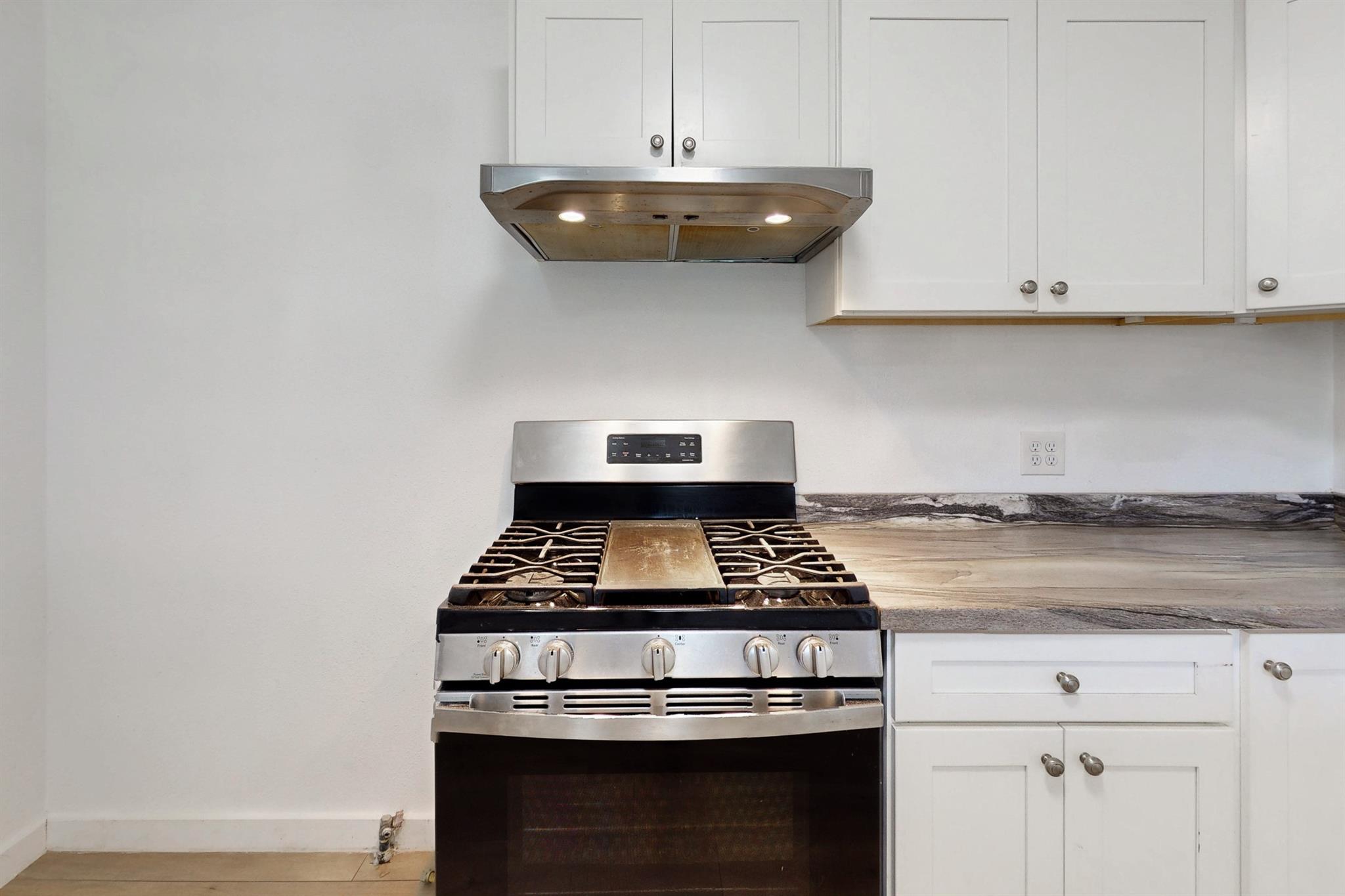 Kitchen range and oven beneath a stainless hood, framed by white cabinets and a wood-look countertop.