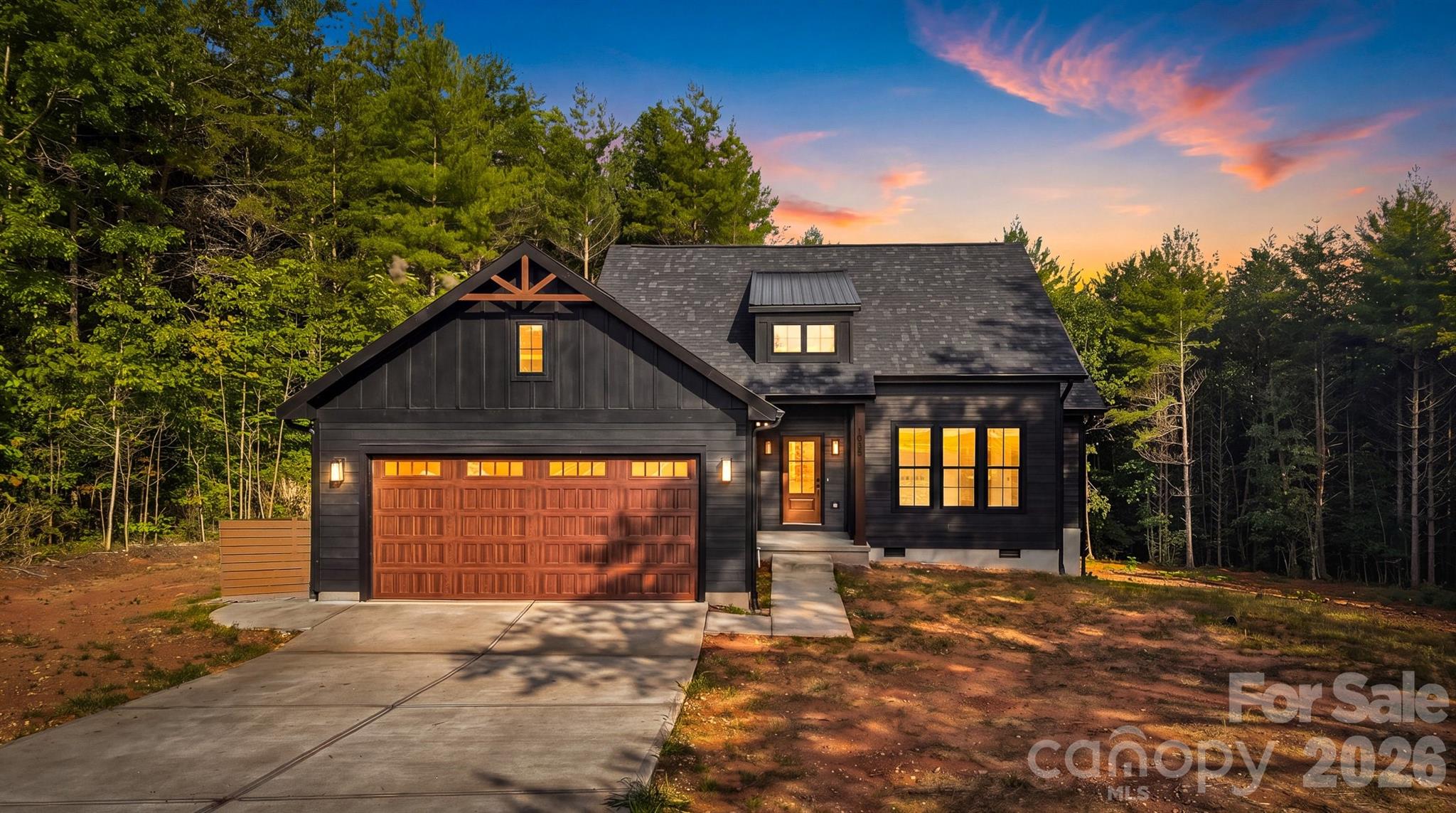 Dark modern farmhouse with a timber-trimmed gable and lit windows among trees at sunset.
