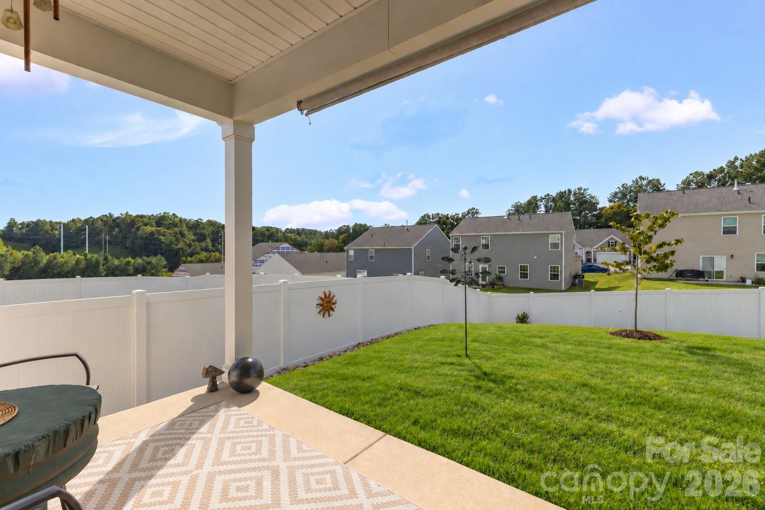 Covered patio overlooking a fenced green backyard with a young tree and neighboring homes beyond.