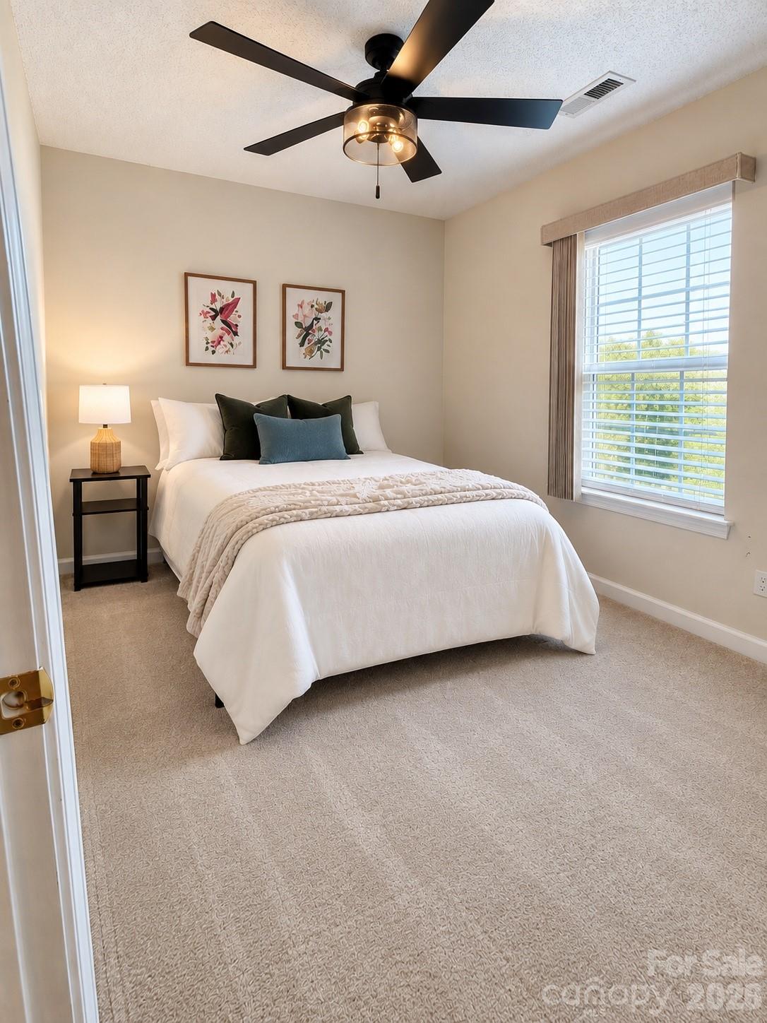 Staged bedroom with a white bed, black ceiling fan, floral prints, and a large window.