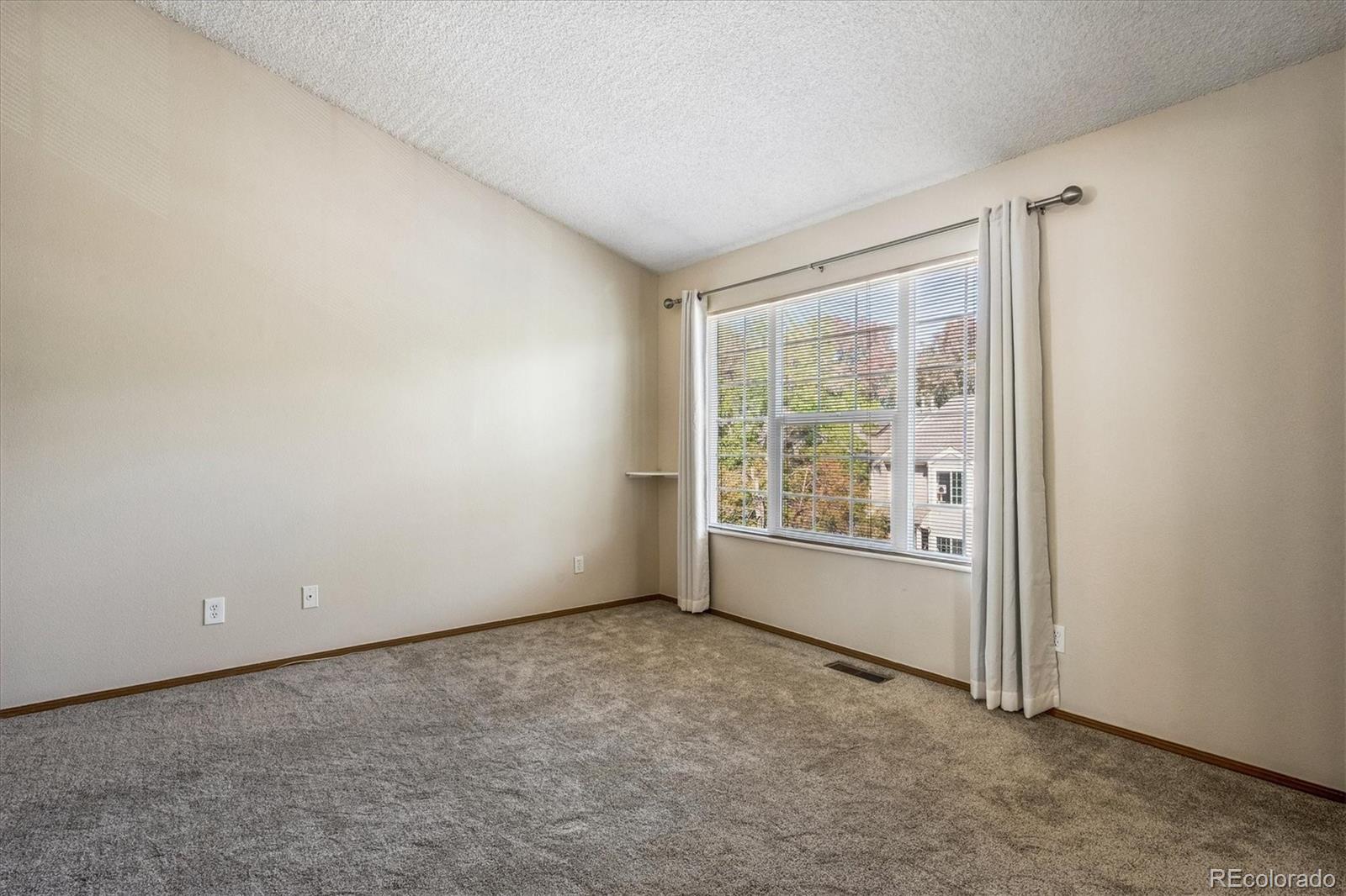 Vacant carpeted room with a large window, blinds, and white curtains.