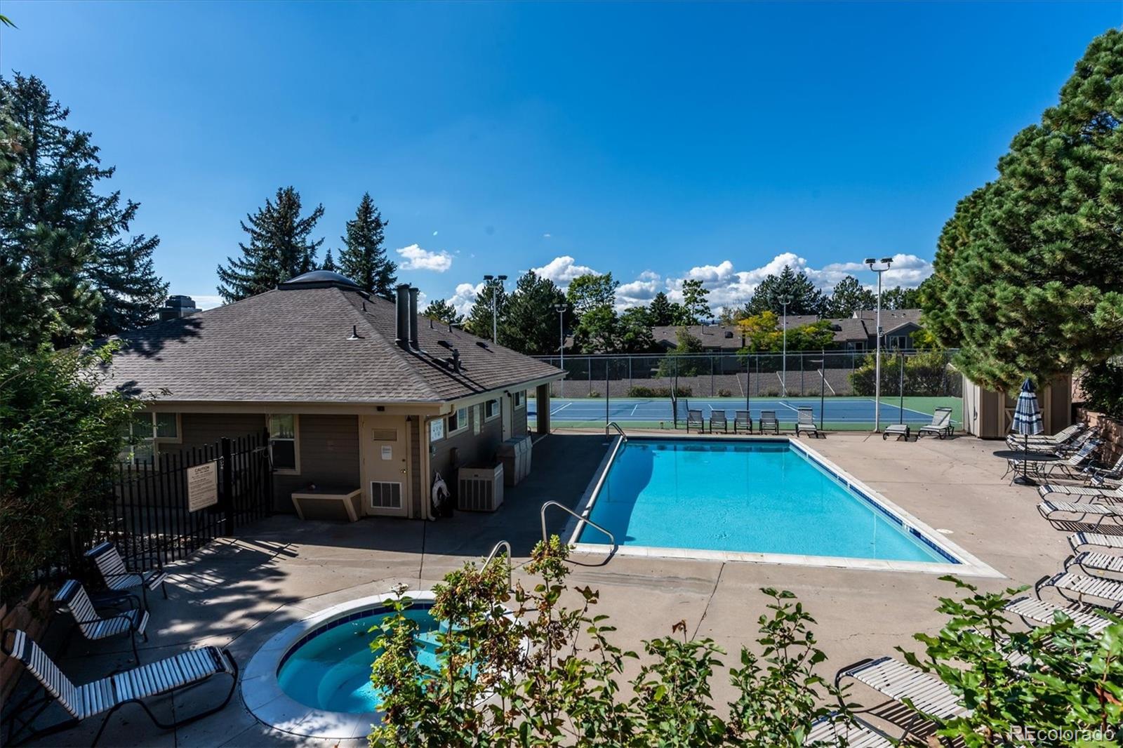 Community pool and circular hot tub beside a fenced tennis court and poolside lounge chairs.