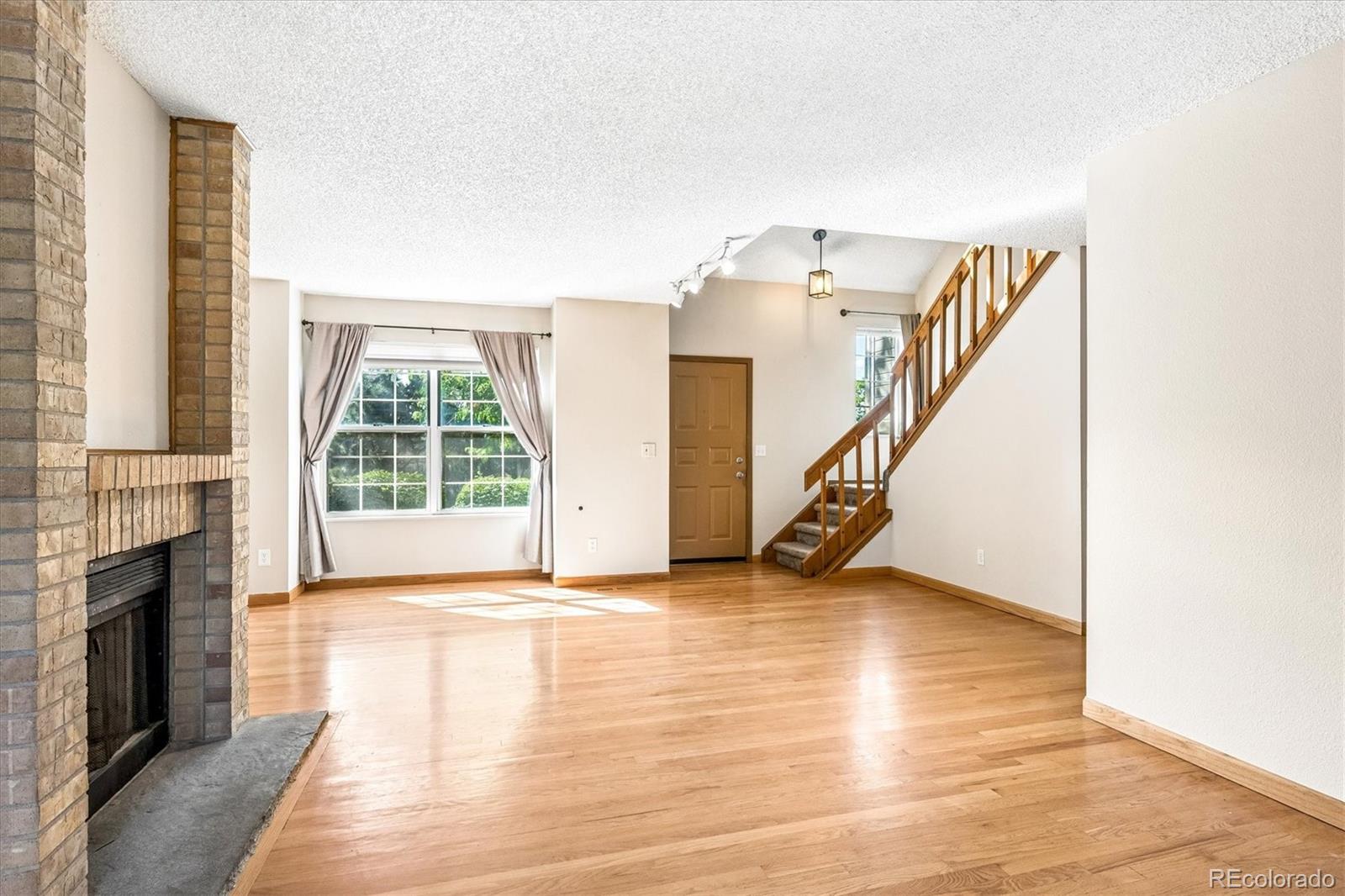 Vacant living room with hardwood floors, brick fireplace, and wooden staircase.